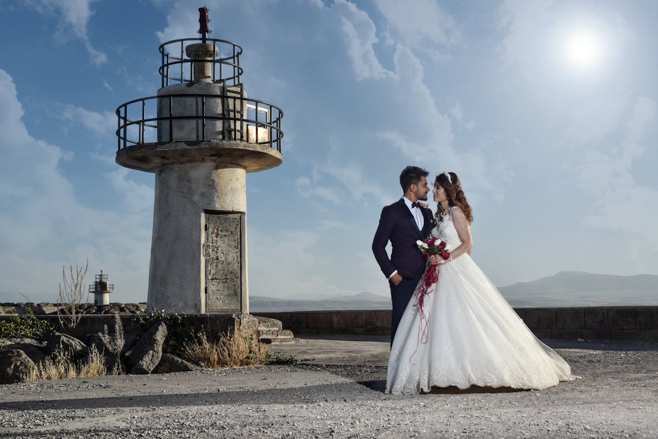 A romantic wedding portrait of a couple by a rustic lighthouse under a clear blue sky.